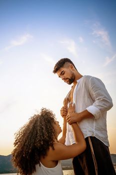 Romance scene of handsome bearded guy holding his kneeling girlfriend by the wrists looking in eyes at sunset in nature. Submissive young afro american curly woman in love in adoration of her man