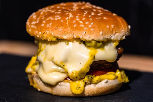 Homemade beef cheeseburger with melting cheese, tomatoes, onions, pickles isolated on a black stone plate.