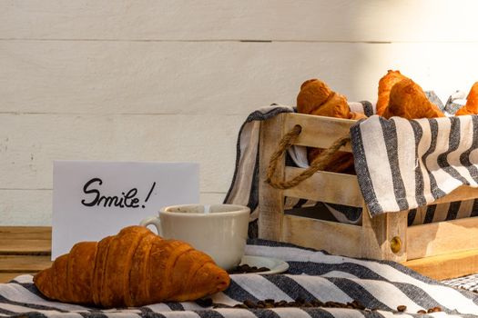 Coffee cup and buttered fresh French croissant on wooden crate. Food and breakfast concept. Morning message “smile” on white board