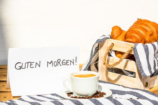 Coffee cup and buttered fresh French croissant on wooden crate. Food and breakfast concept. Morning message in German meaning “good morning” on white board
