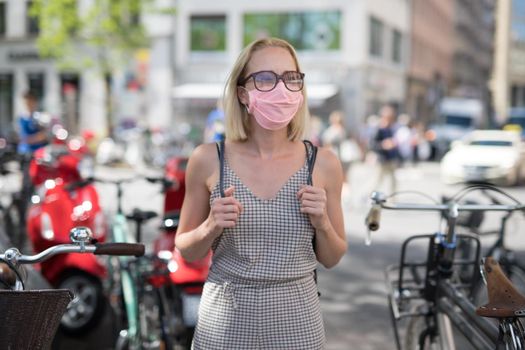 Portrait of casual yound woman walking on the street wearing protective mask as protection against covid-19 virus. Incidental people on the background.