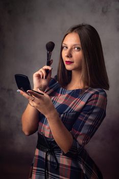 The girl powders her face with a brush while looking in the mirror, the girl was distracted and looked into the frame, studio photography on a gray background