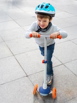 Toddler in helmet is riding scooter on parking lot with parked bicycles. Urban vehicle for active children. Leisure activity for boys and girls.