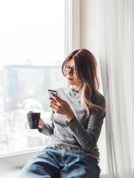 Thoughtful woman with eyeglasses and cup of hot coffee looks at her smartphone. Information in online media.
