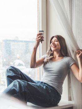 Woman with curly hair poses for selfie on windowsill. Pretty woman makes self-portrait on smartphone. Winter morning at home.