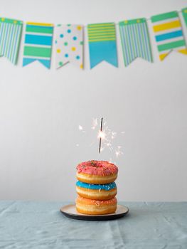 Birthday donuts cake with bengal light or sparkler. Stack of delicious doughnuts over table with party flag garland on background. Vertical. Copy space
