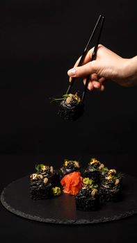 Hand with chopsticks holds a sushi. Custom sushi roll with black rice, crab meat, avocado, smoked salmon mousse, oar caviar, masago, shrimp cocktail, edible gold leaf, ginger, wasabi on black table