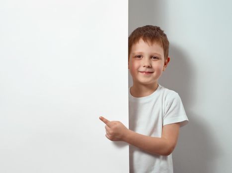 Four-year-old boy shows finger on blank white sheet. Happy child in white t-shirt over white background with copy space for message, mock up