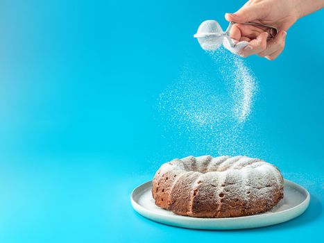 Woman's hand sprinkling icing sugar over fresh home made bundt cake. Powder sugar falls on fresh perfect bunt cake over blue background. Copy space for text. Ideas and recipes for breakfast or dessert