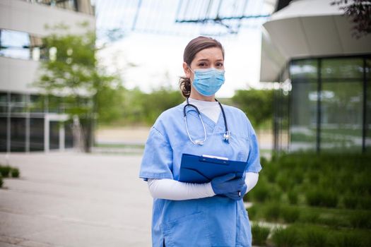 Young female EMS key worker doctor in front of healthcare ICU facility,wearing protective face mask holding medical patient lab health check form,COVID-19 pandemic outbreak crisis PPE shortage in UK