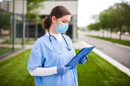 Female UK NHS doctor holding blue clipboard standing outside hospital clinic street entrance,frontline key medical worker studying patient card form,modern care facility nursing home complex exterior