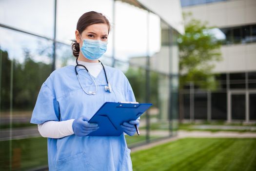 Portrait of tired exhausted female caucasian NHS UK EMS doctor in front of clinic or hospital,wearing uniform & face mask,Coronavirus COVID-19 virus disease,global pandemic outbreak,testing & analysis