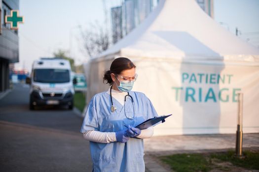 Female key doctor checking patient report form medical card,standing outside clinic exterior triage tent,Coronavirus COVID-19 illness diagnosis test,emergency due to global pandemic crisis,NHS UK EMS