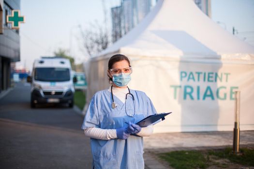 Portrait of serious female key front line worker in blue PPE uniform,standing outside EMS hospital or ICU clinic facility entrance,UK COVID-19 drive through testing site,rt-PCR Coronavirus diagnostic