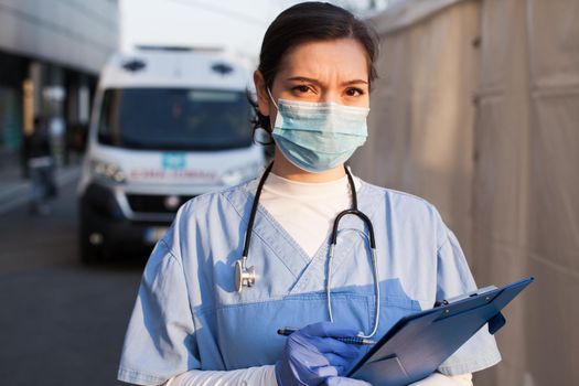 Young female NHS UK EMS doctor in front of healthcare ICU facility,wearing protective face mask holding medical patient health check form,Coronavirus COVID-19 pandemic outbreak crisis PPE shortage