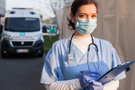 Worried & anxious UK NHS EMS doctor,holding patient report form folder,standing in front of ambulance car,tired & exhausted from working long shifts,outdoor PPE portrait,Coronavirus COVID-19 pandemic