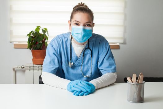 Young female caucasian General Practitioner sitting by her desk in office,wearing blue uniform,protective gloves & face mask,virtual tele visit via video call,GP doctor on demand telemedicine concept