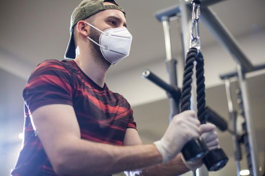 Young caucasian man working out on lat pull-down machine in re-open US gym,wearing protective latex gloves & face mask,activity & staying fit under COVID-19 pandemic lockdown,prevention of Coronavirus
