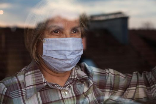 Elderly caucasian woman wearing handmade protective face mask,nursing care home,looking outside the window with sadness in her eyes,self isolation due to the global COVID-19 Coronavirus pandemic,hope