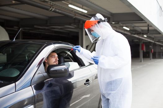 Drive-thru COVID-19 test site,medical staff wearing PPE and face shield performing nasal swabbing,young male driver patient waiting sitting in automobile while worker takes specimen through car window