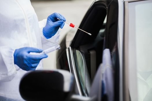 Medical worker in N95 PPE performing nasal & throat swab on person in vehicle through car window,COVID-19 UK NHS mobile testing centre drive thru facility,hands closeup in blue gloves holding test kit