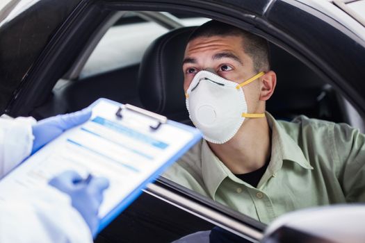 Male driver wearing protective N95 face mask sitting by left drive wheel in UK drive-thru COVID-19 test centre,answering health check up questions,medical worker ticking off symptoms on clipboard form