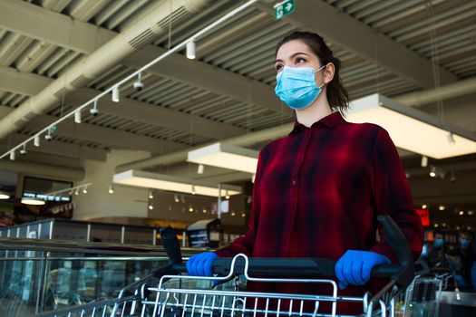 Young caucasian woman pushing shopping cart in supermarket, wearing protective blue surgical face mask and latex gloves, health and safety measurements in public due to Coronavirus COVID-19 pandemic