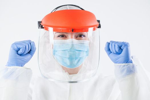 Healthcare professional in personal protective equipment, wearing surgical gloves, face shield, white protective hazmat biohazard suit, clenched fists raised in air and smile,happy and victorious