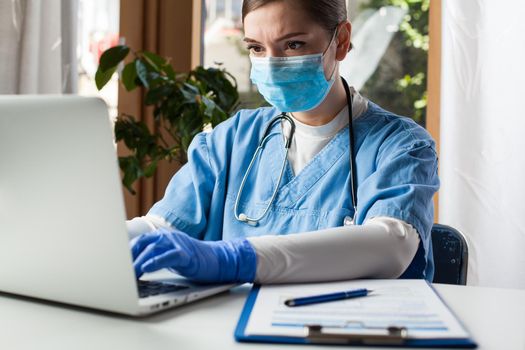 Young caucasian female doctor working on laptop computer in office,wearing protective glove and face mask,Electronic Medical Records EMR concept,remote tele health medicine during Coronavirus pandemic