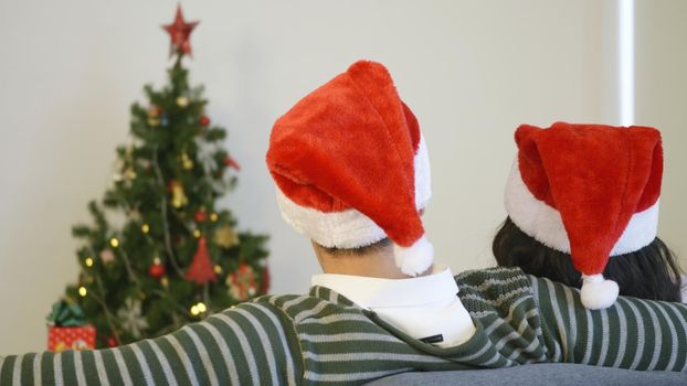 Happy Asian beautiful young family couple husband and wife sitting on sofa wearing a santa hat in front of fireplace at warm home Christmas winter hugging living room, back view,