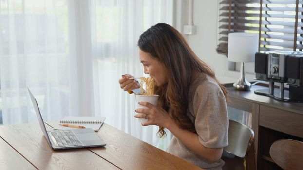 Asian business woman eating instant noodles while working on laptop computer at home office, Happy beautiful young female sitting on desk work overtime doing deadline project, late time business