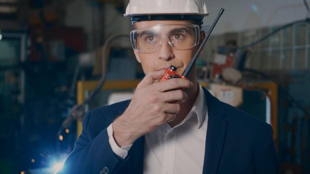 Professional confident engineer businessman in suits safety hats and goggles stand holding walkie-talkie in an heavy industrial facility manufacturing with a steel welding from robot with fire spark