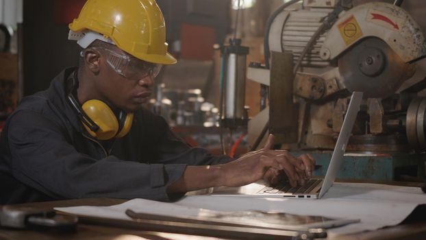 American industrial black young worker man with yellow helmet and ear protection typing keyboard of laptop computer in front machine, Confident engineer at work in the industry factory