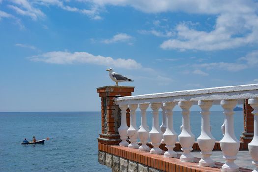 A large Seagull sits on a stone balcony against a blue sea and a boat .