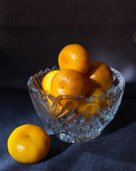 Yellow tangerines in a crystal vase on a dark background.