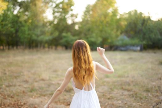 happy red-haired woman in white dress runs on dry grass in nature near trees. High quality photo