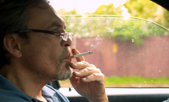 Close up portrait of handsome senior man driving a car and smoking. Shooting inside the car