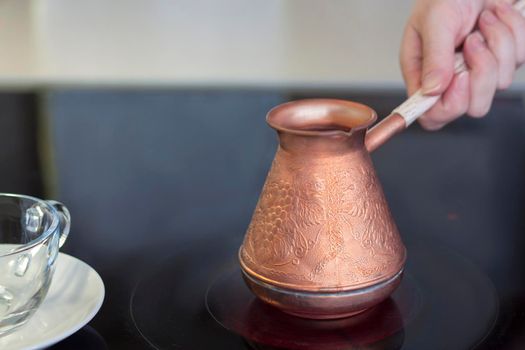 A copper turk for brewing coffee stands on a glass-ceramic plate. A woman's hand holds the handle of a turk. Make coffee in a turk. Aromatic coffee.