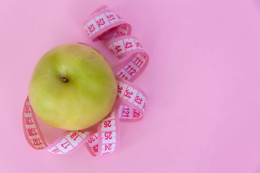 A green apple and a centimeter measuring tape on a pink background. Sports, sports nutrition, healthy eating, diet. Space for the text. Exercise, lose weight.