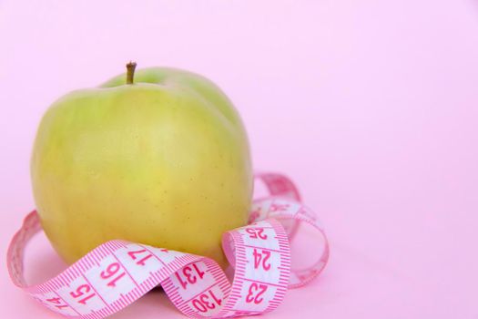 A green apple and a centimeter measuring tape on a pink background. Sports, sports nutrition, healthy eating, diet. Space for the text. Exercise, lose weight.