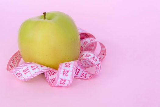 A green apple and a centimeter measuring tape on a pink background. Sports, sports nutrition, healthy eating, diet. Space for the text. Exercise, lose weight. 