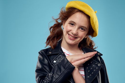 A fashionable girl in a leather jacket and a yellow hat on a blue background gestures with her hands emotions. High quality photo