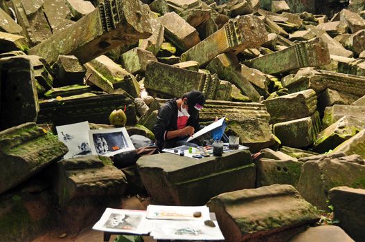 ANGKOR, CAMBODIA - AUG 28, 2013: Man sitting on stone remnants of building in complex of Angkor Wat and drawing paintings. Painter on ruins of temple