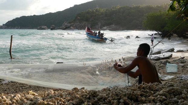KOH PHANGAN, THAILAND - 11 JANUARY 2019 Ethnic fisherman hanging bait on net. Side view of ethnic shirtless man hanging small fish on net while sitting on shore near sea.