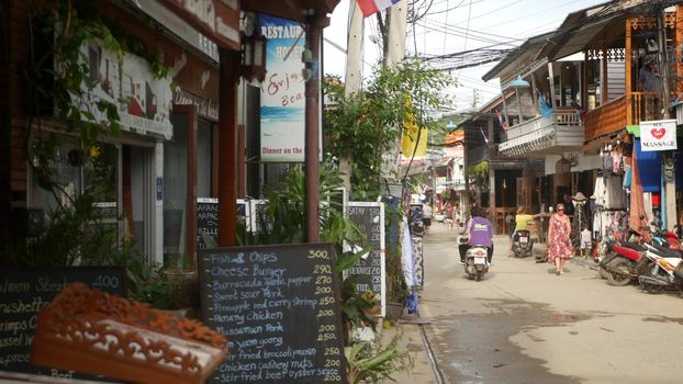 SAMUI ISLAND, THAILAND - MAY 27, 2019: Typical touristic street in Fisherman village with souvenir stores. View of calm lane of city in Asia with touristic shops in daytime. Thai people on motorbikes