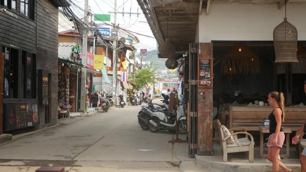 SAMUI ISLAND, THAILAND - MAY 27, 2019: Typical touristic street in Fisherman village with souvenir stores. View of calm lane of city in Asia with touristic shops in daytime. Thai people on motorbikes