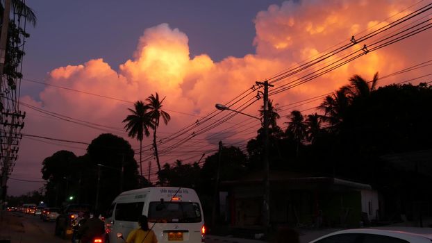 KOH SAMUI ISLAND, THAILAND - 10 JULY 2019: City road with urban transport moving. Back view of cars and motorbikes moving on busy road under orange clouds in sunset tropical exotic evening.