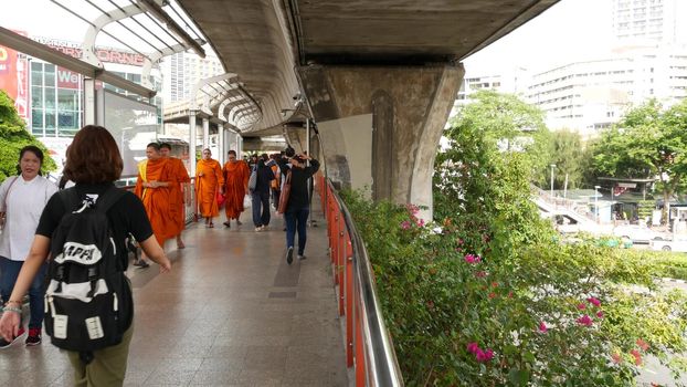 BANGKOK, THAILAND - 10 JULY, 2019: Buddhist monks walking on crowded path. Group of Buddhist monks in traditional orange robes walking on pedestrian path near crowd of modern people on street.