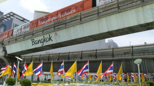 BANGKOK, THAILAND - 11 JULY, 2019: Pedestrians walking on the bridge near MBK and Siam Square under BTS train line. People in festive modern city decorated with waving national flags and royal symbol.