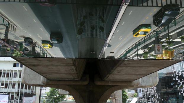 BANGKOK, THAILAND - 11 JULY, 2019 Intersection on busy city street. People on motorcycles and cars riding, pedestrian bridge. Vehicles reflected upside down in mirror. City life perspective reflection